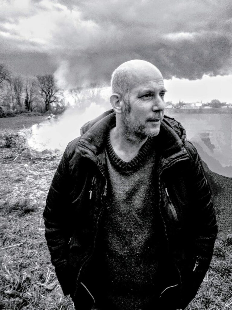 Stefan Jäkel in dunkler Jacke und Strickpullover am Ufer eines Gewässers.