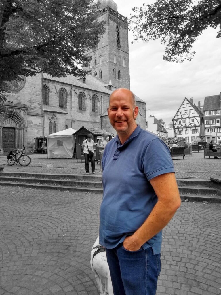 Stefan Jäkel im blauen Poloshirt auf einem Kopfsteinpflasterplatz; im Hintergrund ein weißer Hund, eine Kirche und Fachwerkhäuser.